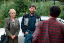 Two adults stand by a silver car talking to a person in a red plaid shirt (back to camera) in an outdoor setting with greenery in the background.