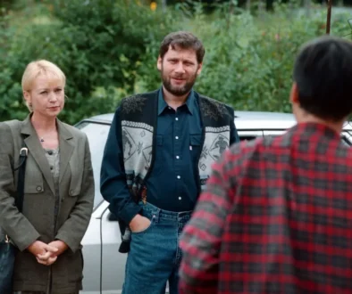 Two adults stand by a silver car talking to a person in a red plaid shirt (back to camera) in an outdoor setting with greenery in the background.