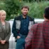 Two adults stand by a silver car talking to a person in a red plaid shirt (back to camera) in an outdoor setting with greenery in the background.