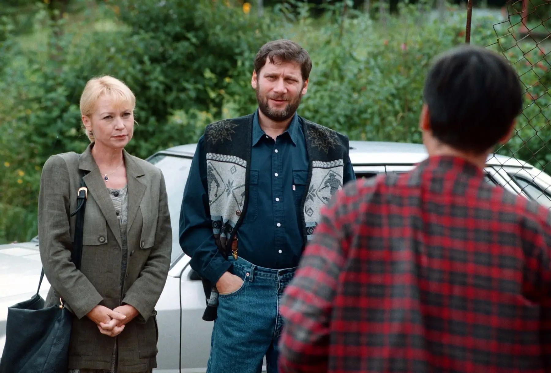 Two adults stand by a silver car talking to a person in a red plaid shirt (back to camera) in an outdoor setting with greenery in the background.