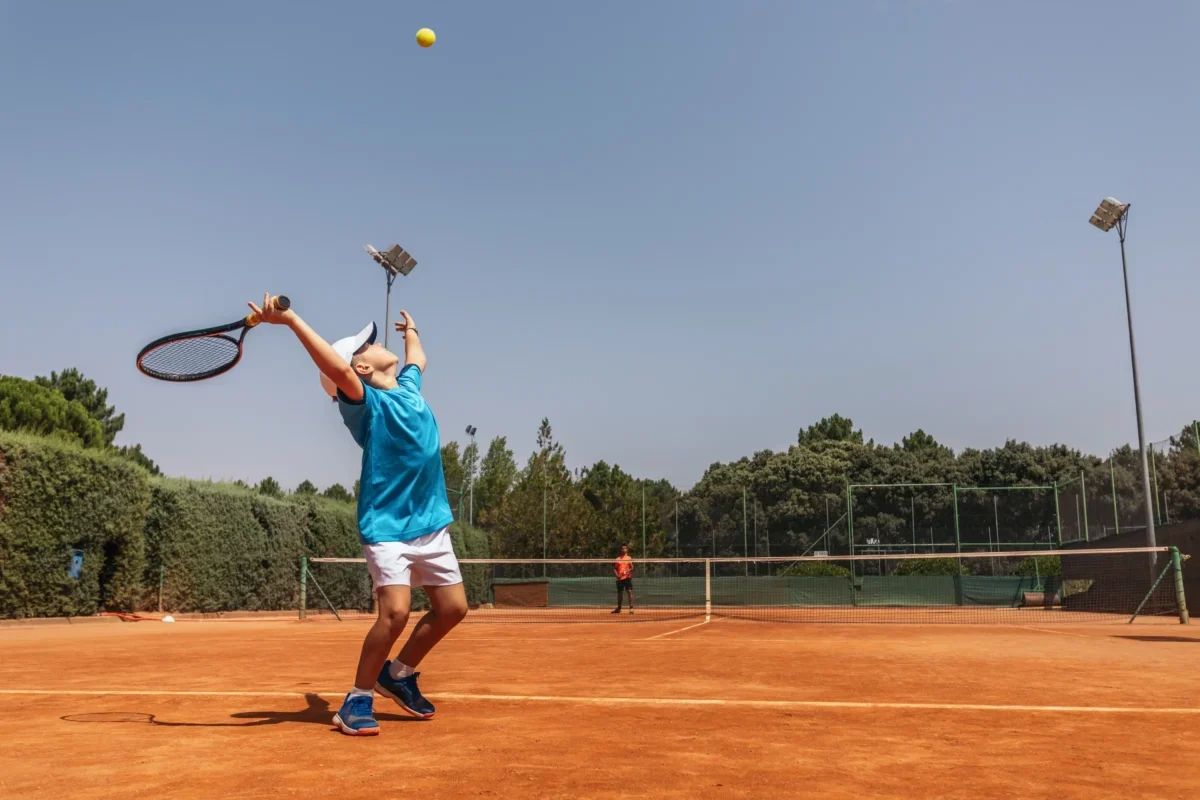 Tennis player in a blue shirt and white cap serving on a clay court, ball midair above the racket on a sunny day.