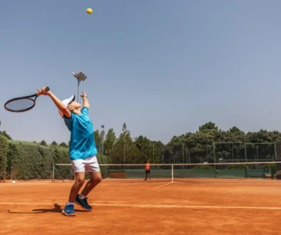 Tennis player in a blue shirt and white cap serving on a clay court, ball midair above the racket on a sunny day.