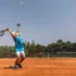 Tennis player in a blue shirt and white cap serving on a clay court, ball midair above the racket on a sunny day.