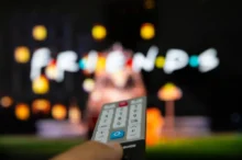Close-up of a TV remote held toward a blurred colorful screen in the background.