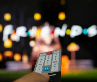 Close-up of a TV remote held toward a blurred colorful screen in the background.