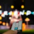 Close-up of a TV remote held toward a blurred colorful screen in the background.