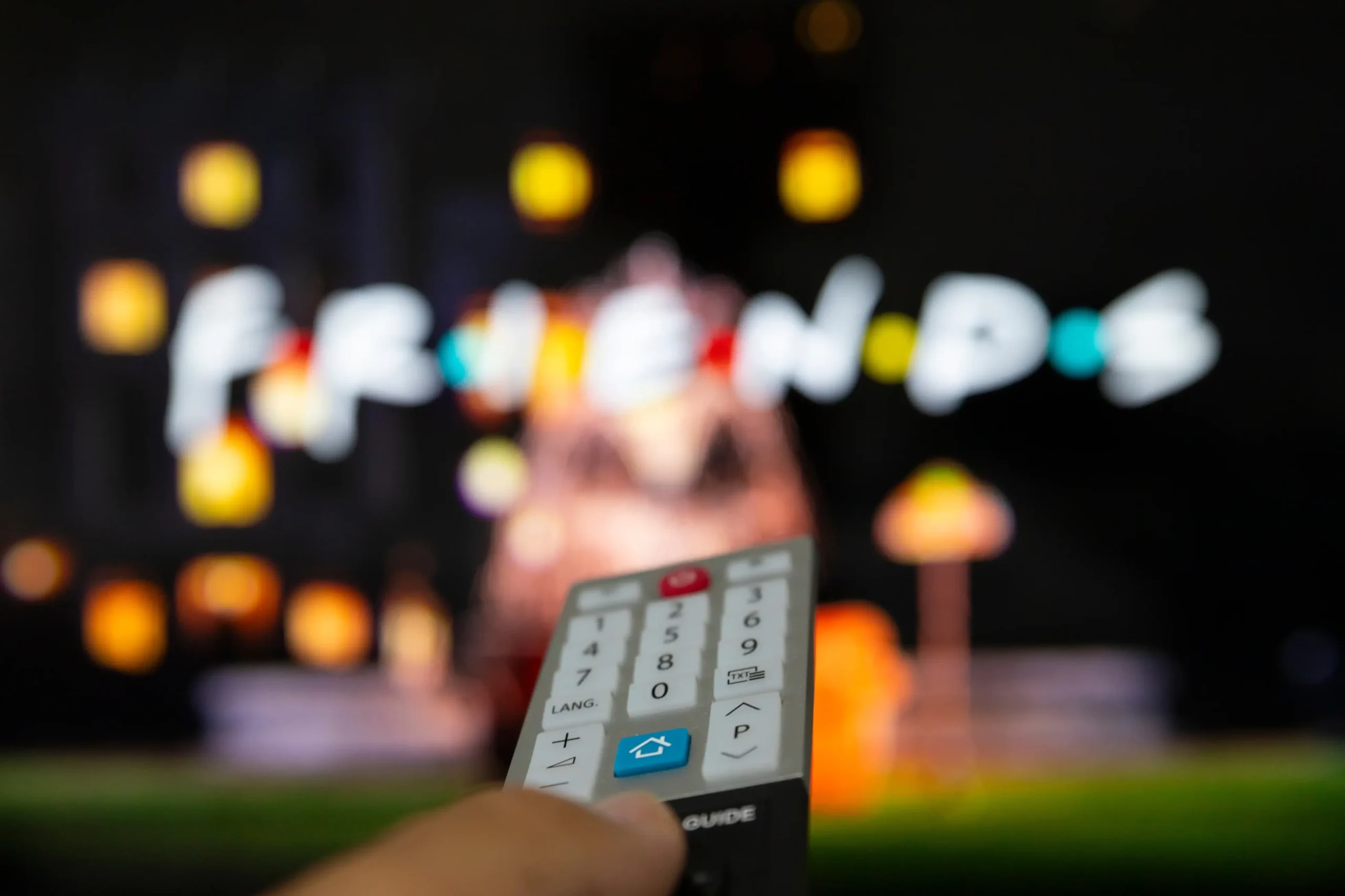 Close-up of a TV remote held toward a blurred colorful screen in the background.