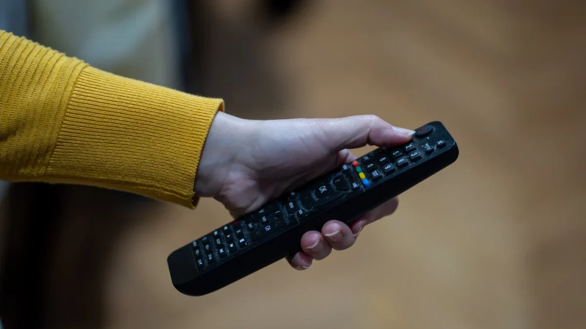 Close-up of a hand with a yellow-sleeved sweater gripping a black TV remote, showing colorful buttons