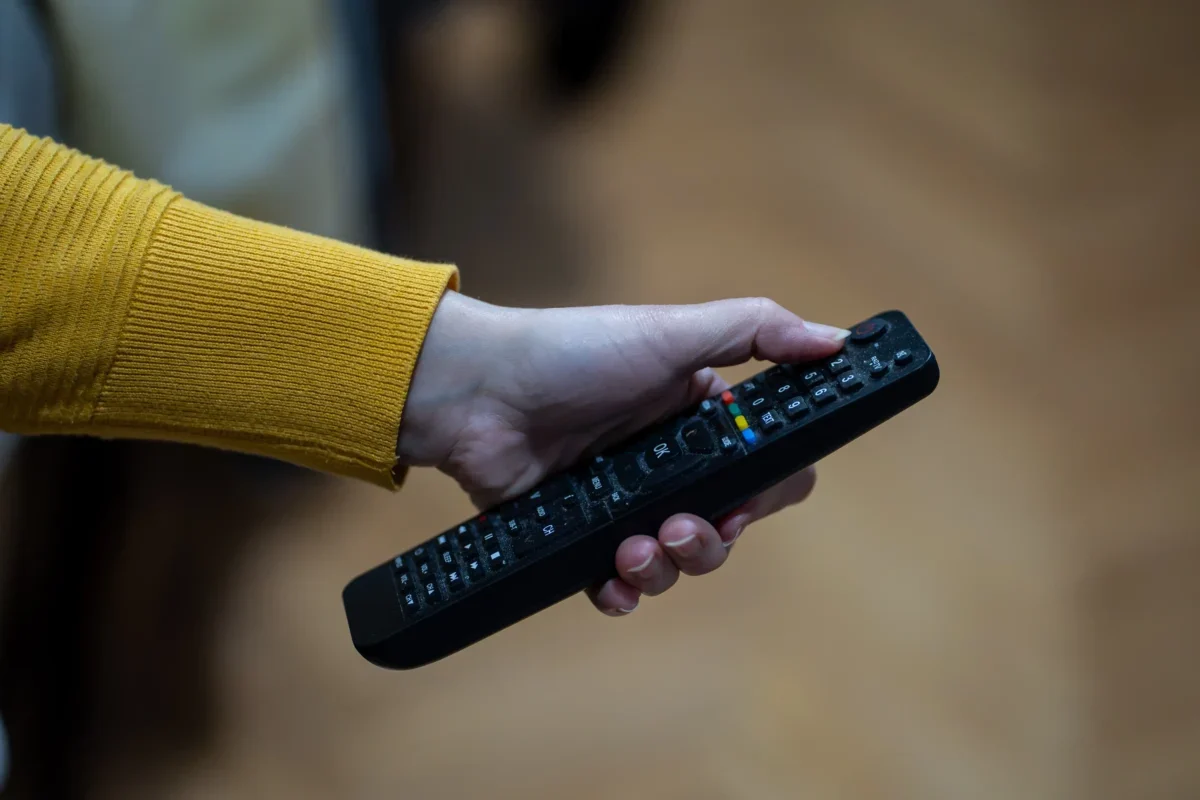 Close-up of a hand with a yellow-sleeved sweater gripping a black TV remote, showing colorful buttons