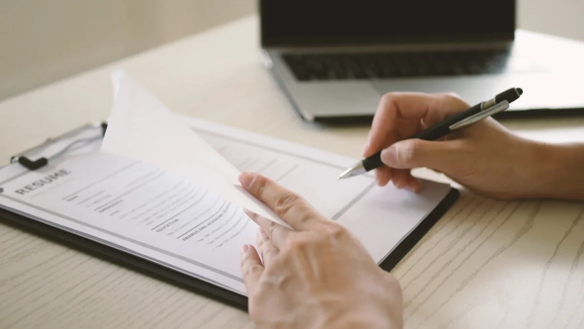 Person signing a resume on a clipboard with a pen, laptop in the background