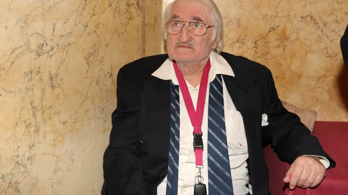 Older man with white hair and glasses wearing a black blazer, white shirt, striped tie, and a pink lanyard, seated against a marble wall.