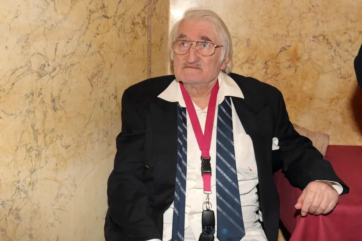 Older man with white hair and glasses wearing a black blazer, white shirt, striped tie, and a pink lanyard, seated against a marble wall.
