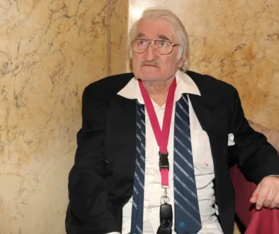 Older man with white hair and glasses wearing a black blazer, white shirt, striped tie, and a pink lanyard, seated against a marble wall.