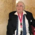 Older man with white hair and glasses wearing a black blazer, white shirt, striped tie, and a pink lanyard, seated against a marble wall.
