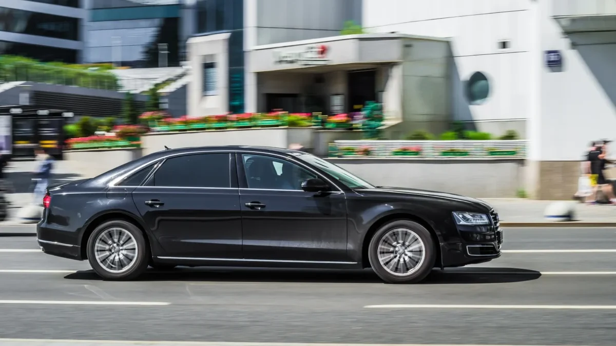 Black luxury sedan driving past urban buildings in motion blur, street scene with colorful planters and pedestrians in the background
