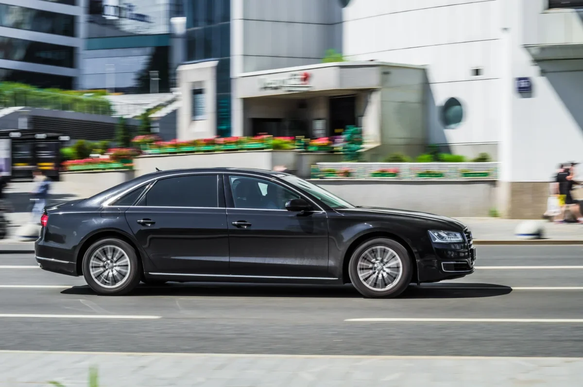 Black luxury sedan driving past urban buildings in motion blur, street scene with colorful planters and pedestrians in the background