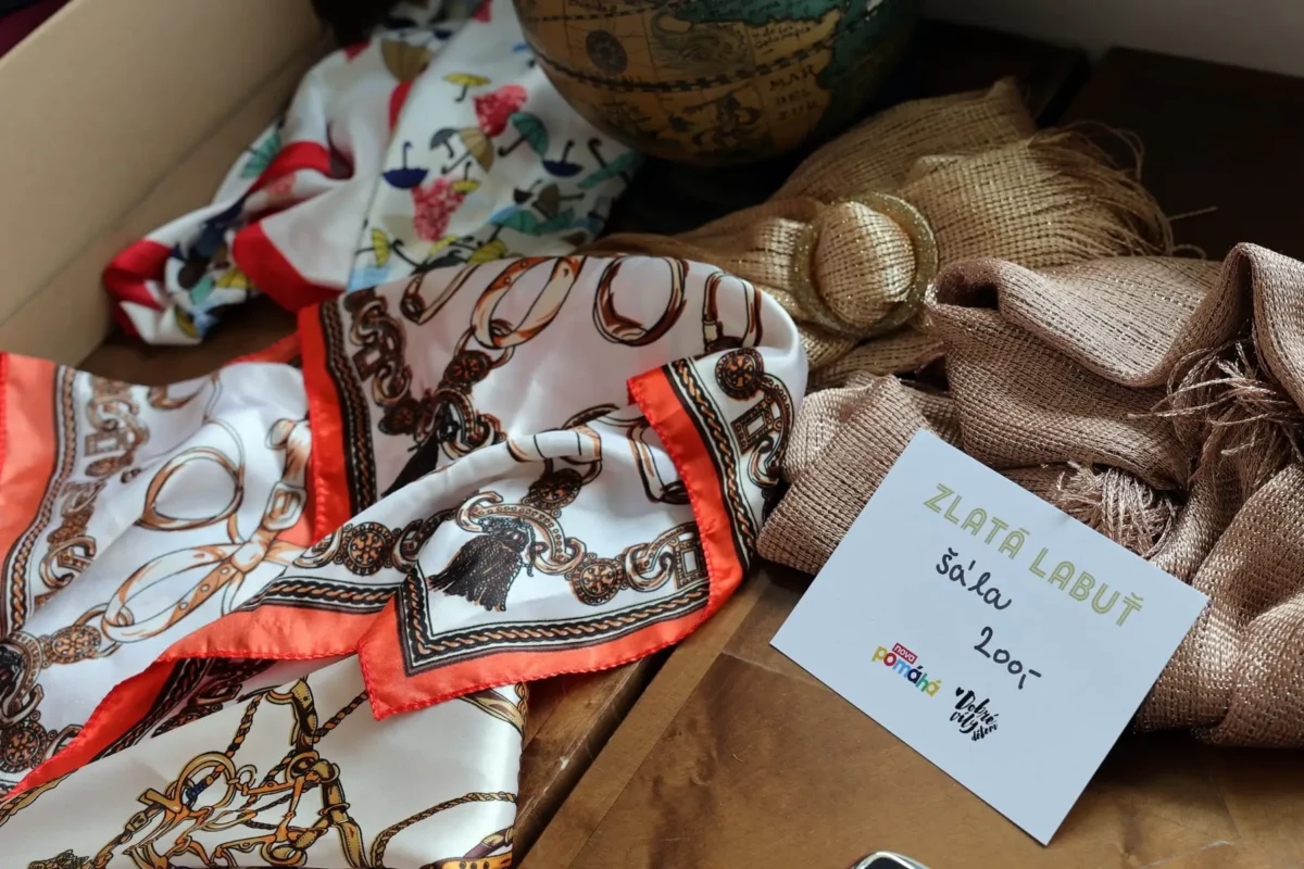profimedia-0867020395 1 | TVGURU.cz Close-up of a patterned scarf with an orange border, paired with a textured burlap ribbon on a wooden surface, with a globe in the background and a light blue label nearby.