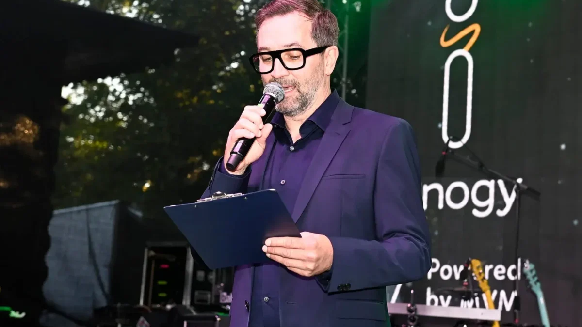 Male presenter in a navy suit speaks into a handheld microphone, holding a clipboard on stage with a banner in the background.