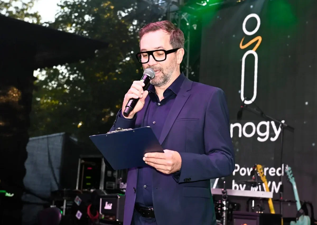 Male presenter in a navy suit speaks into a handheld microphone, holding a clipboard on stage with a banner in the background.