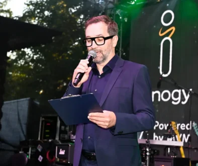 Male presenter in a navy suit speaks into a handheld microphone, holding a clipboard on stage with a banner in the background.