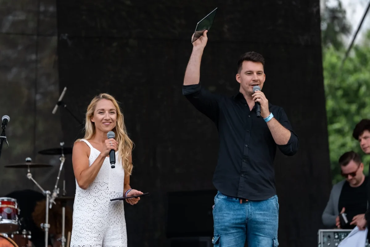 shutterstock_2164858467 1 | TVGURU.cz Two hosts on an outdoor stage: woman in a white dress holds a microphone and tablet, man in a dark shirt raises a tablet while speaking into a mic.