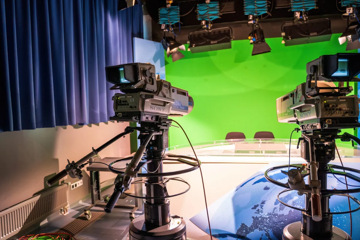 shutterstock_2721484641 1 | TVGURU.cz Two Sony broadcast cameras on tripods in a studio with a green chroma key wall and blue curtains.