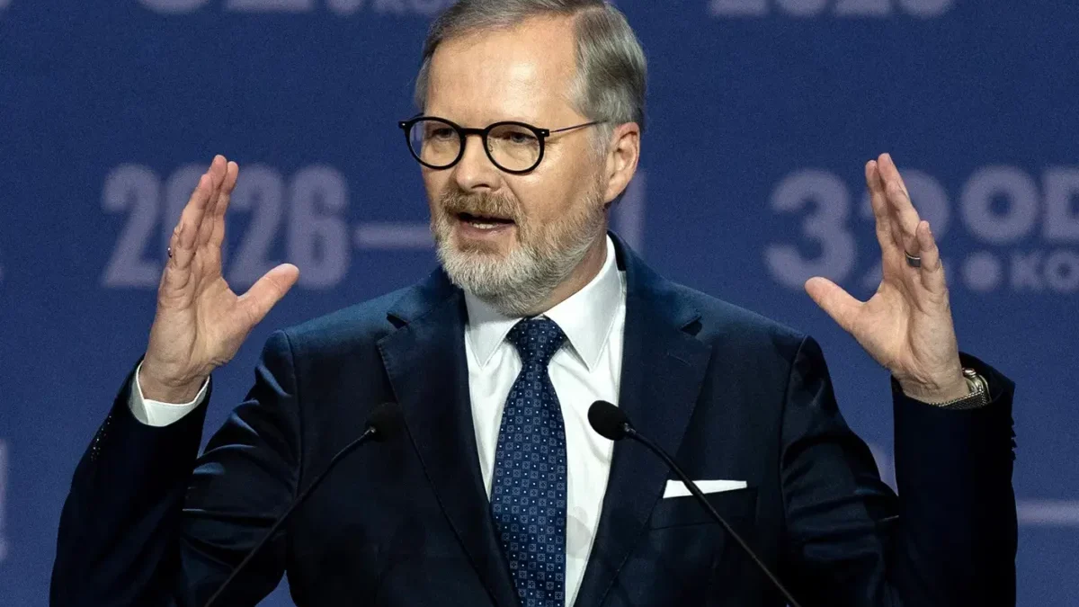 Man in a dark suit and glasses speaking at a podium, hands raised, in front of a blue backdrop with '2026' text behind him.