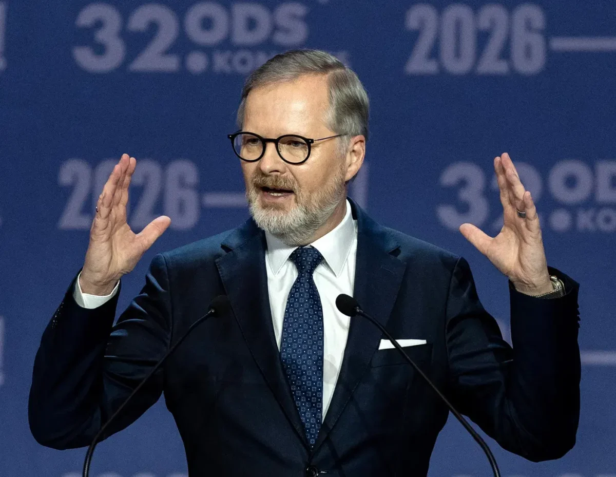 Man in a dark suit and glasses speaking at a podium, hands raised, in front of a blue backdrop with '2026' text behind him.