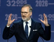 Man in a dark suit and glasses speaking at a podium, hands raised, in front of a blue backdrop with '2026' text behind him.