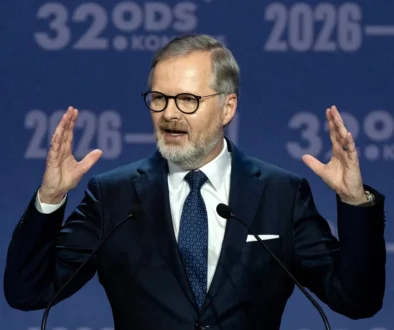 Man in a dark suit and glasses speaking at a podium, hands raised, in front of a blue backdrop with '2026' text behind him.