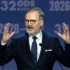 Man in a dark suit and glasses speaking at a podium, hands raised, in front of a blue backdrop with '2026' text behind him.