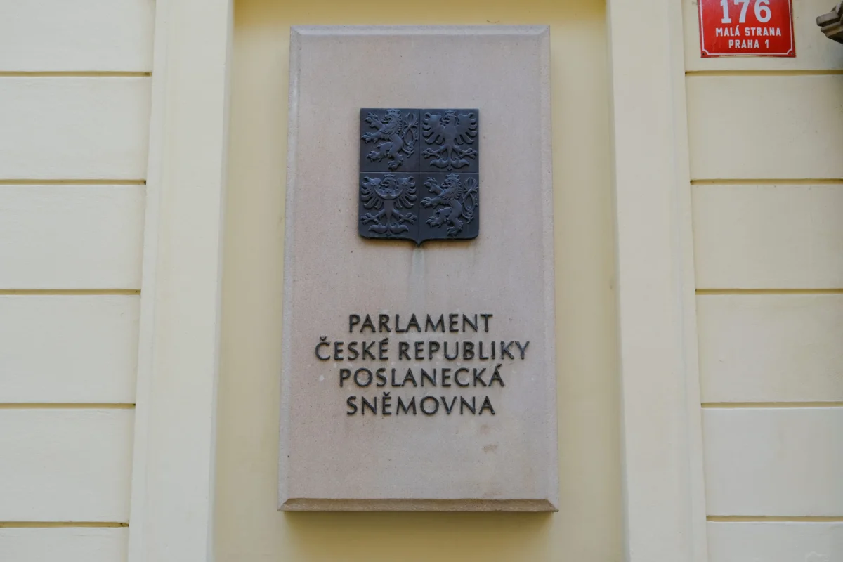 shutterstock_2232179685 1 | TVGURU.cz Stone plaque on a pale yellow wall showing the Czech Parliament emblem and the inscription'Parlament České republiky Poslanecká sněmovna' with a red street sign reading '176 Malá Strana Praha 1'