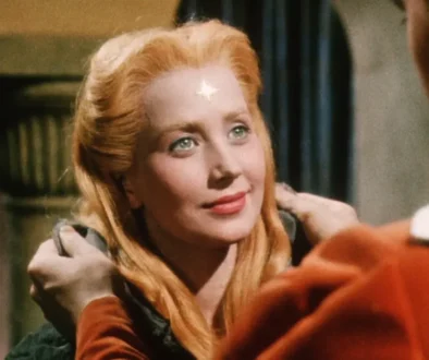 Blonde woman with a star-shaped mark on her forehead gazes up at a person wearing a red uniform, smiling softly.