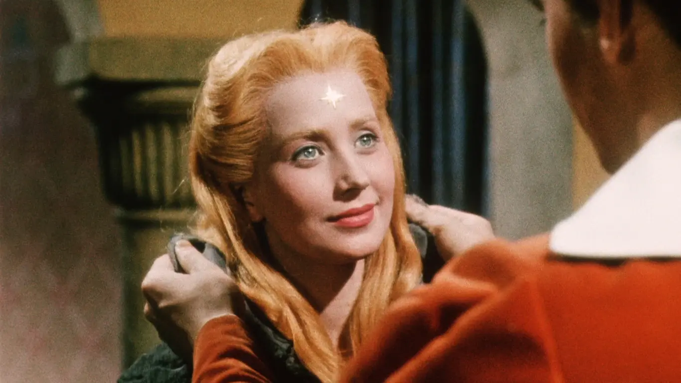 Blonde woman with a star-shaped mark on her forehead gazes up at a person wearing a red uniform, smiling softly.