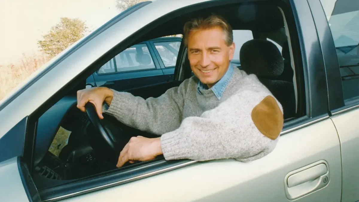 Mž ve svetru sedí v autě a jednu ruku má položenou na volantu, druhou má opřenou o okýnko