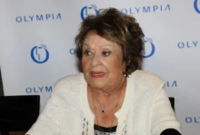 Starší žena s krátkými vlnitými vlasy a v bílé blůze sedí za stolem a dívá se do strany