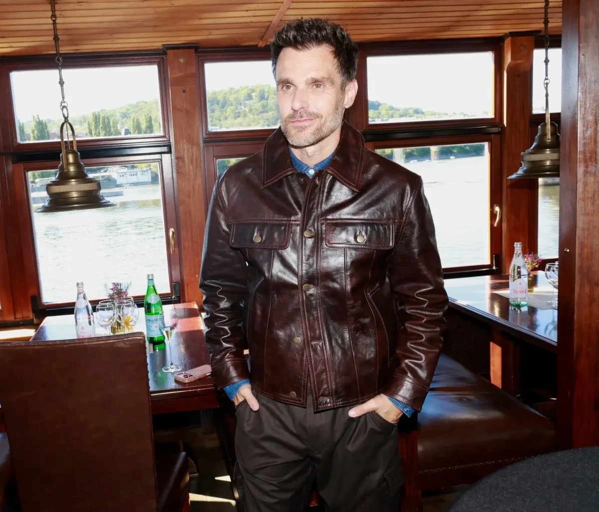 Man in a dark brown leather jacket standing inside a boat restaurant near windows overlooking a river.