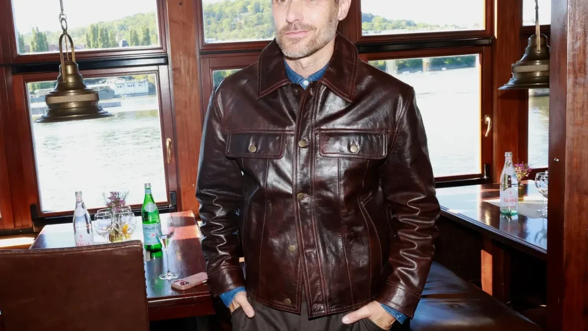 Man in a dark brown leather jacket standing inside a boat restaurant near windows overlooking a river.
