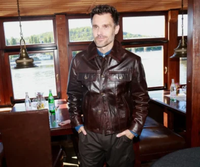 Man in a dark brown leather jacket standing inside a boat restaurant near windows overlooking a river.