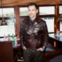 Man in a dark brown leather jacket standing inside a boat restaurant near windows overlooking a river.