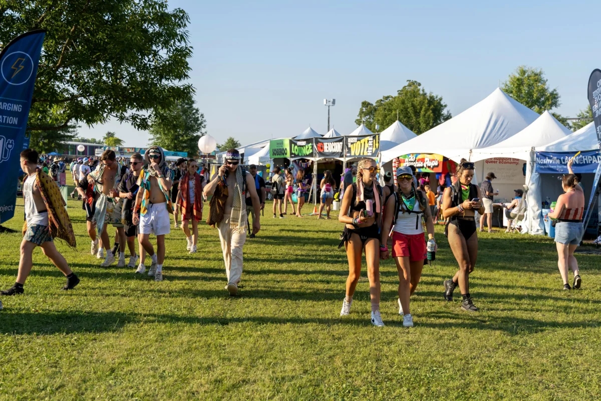 shutterstock_2506073241 2 | TVGURU.cz People in colorful festival attire walking across a sunny grassy field with white event tents and banners in the background.