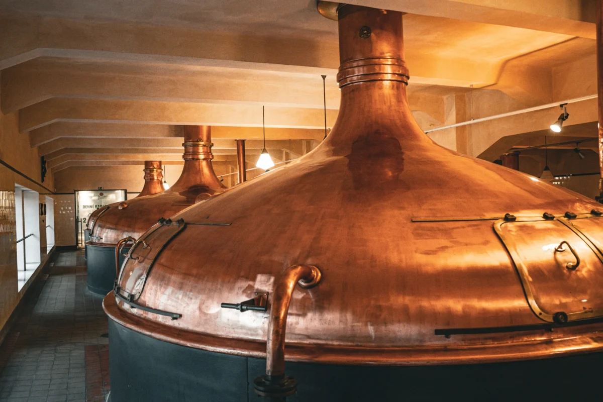 shutterstock_2583008075 1 | TVGURU.cz Copper brewing kettles in a dimly lit brewery with overhead pipes and a tiled floor nearby