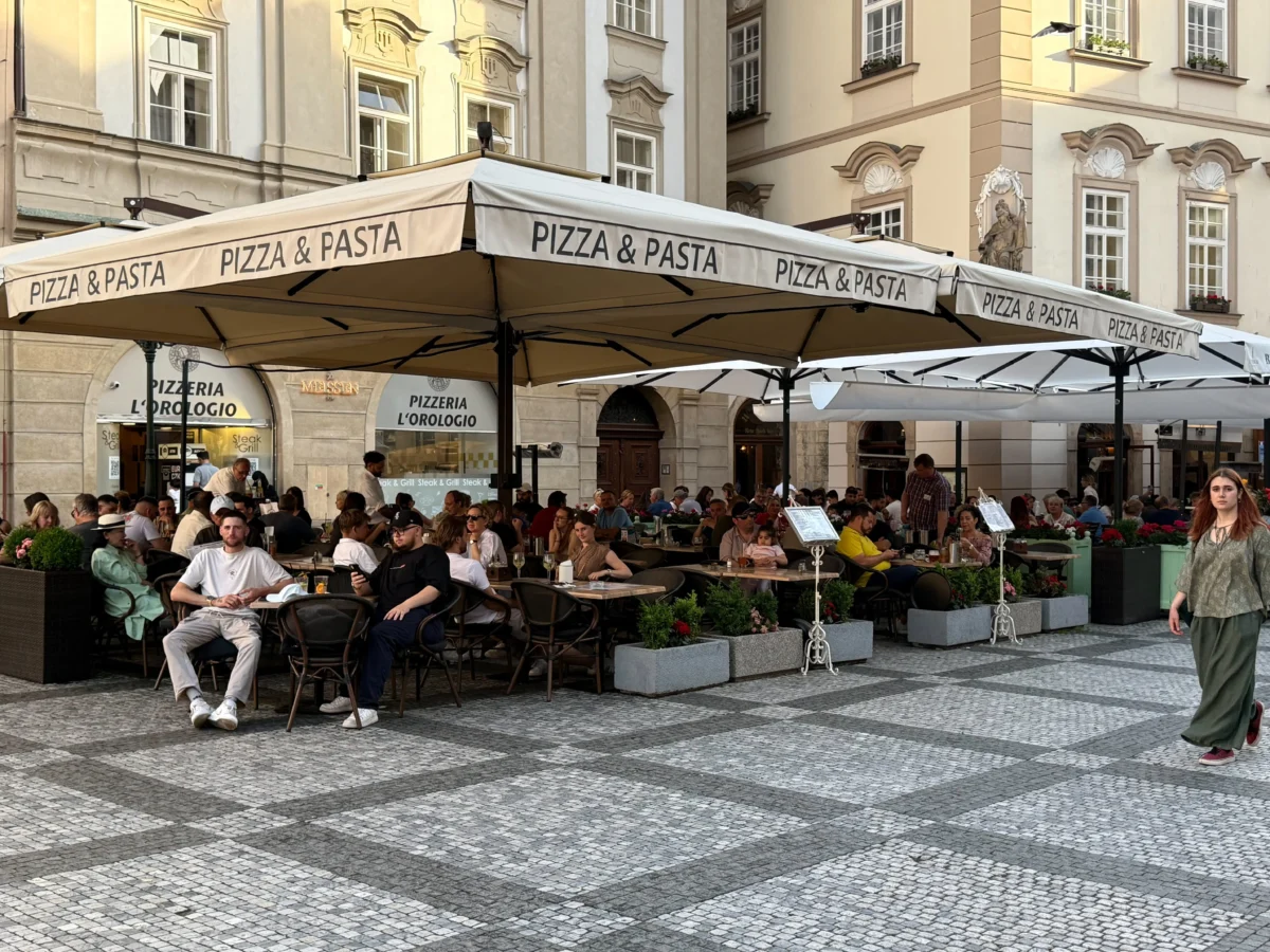 shutterstock_2791321951 2 1 | TVGURU.cz Outdoor pizza and pasta cafe with many people seated under large beige umbrellas in a historic square with cobblestone pavement and pale buildings in the background.