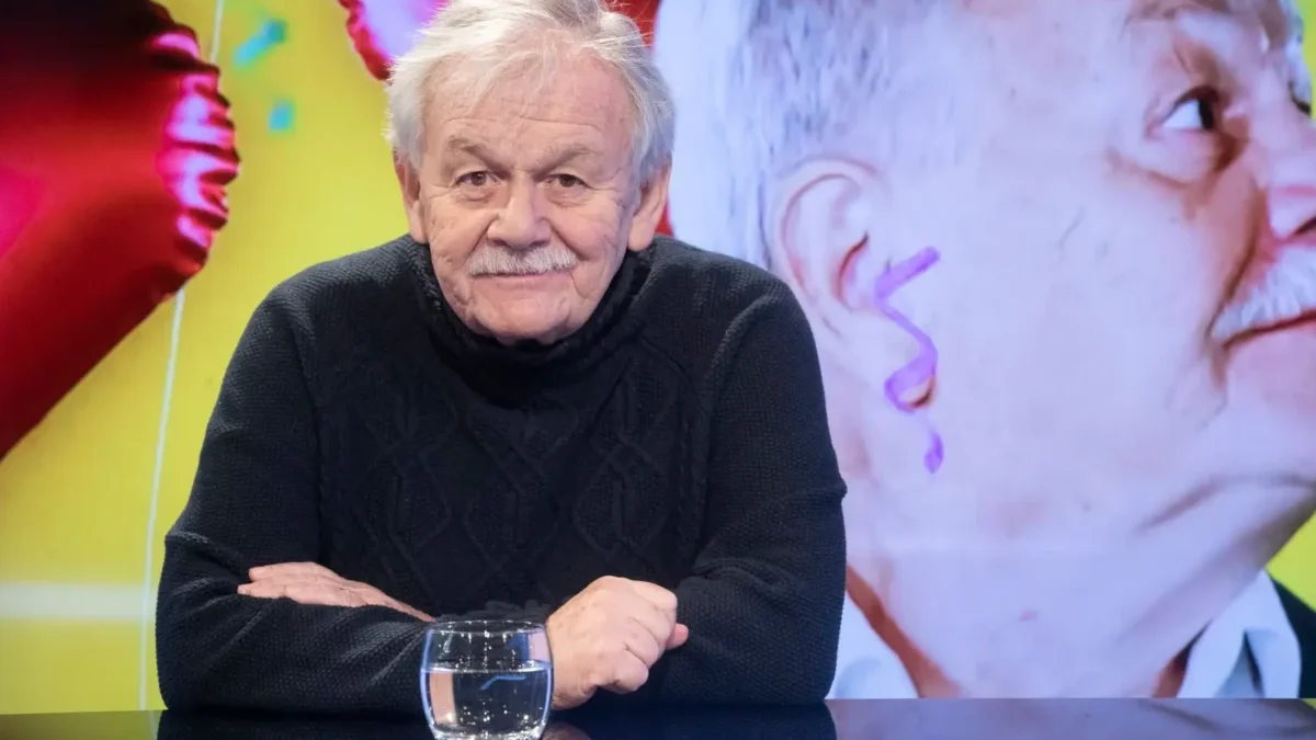 Portrait of an elderly man with gray hair and a mustache, wearing a dark knit sweater, seated at a glass table with a water glass in front of him.