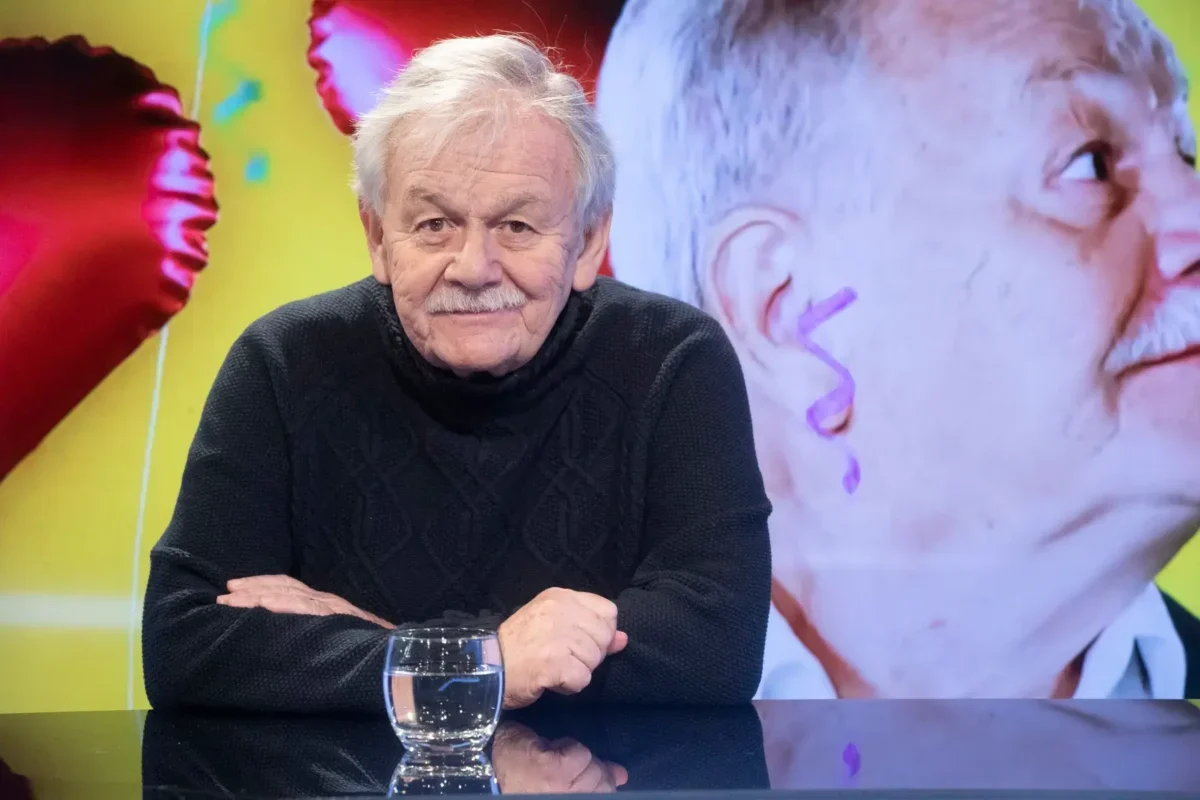 Portrait of an elderly man with gray hair and a mustache, wearing a dark knit sweater, seated at a glass table with a water glass in front of him.