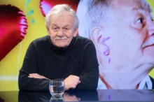 Portrait of an elderly man with gray hair and a mustache, wearing a dark knit sweater, seated at a glass table with a water glass in front of him.