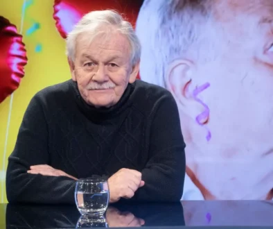 Portrait of an elderly man with gray hair and a mustache, wearing a dark knit sweater, seated at a glass table with a water glass in front of him.