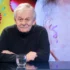 Portrait of an elderly man with gray hair and a mustache, wearing a dark knit sweater, seated at a glass table with a water glass in front of him.