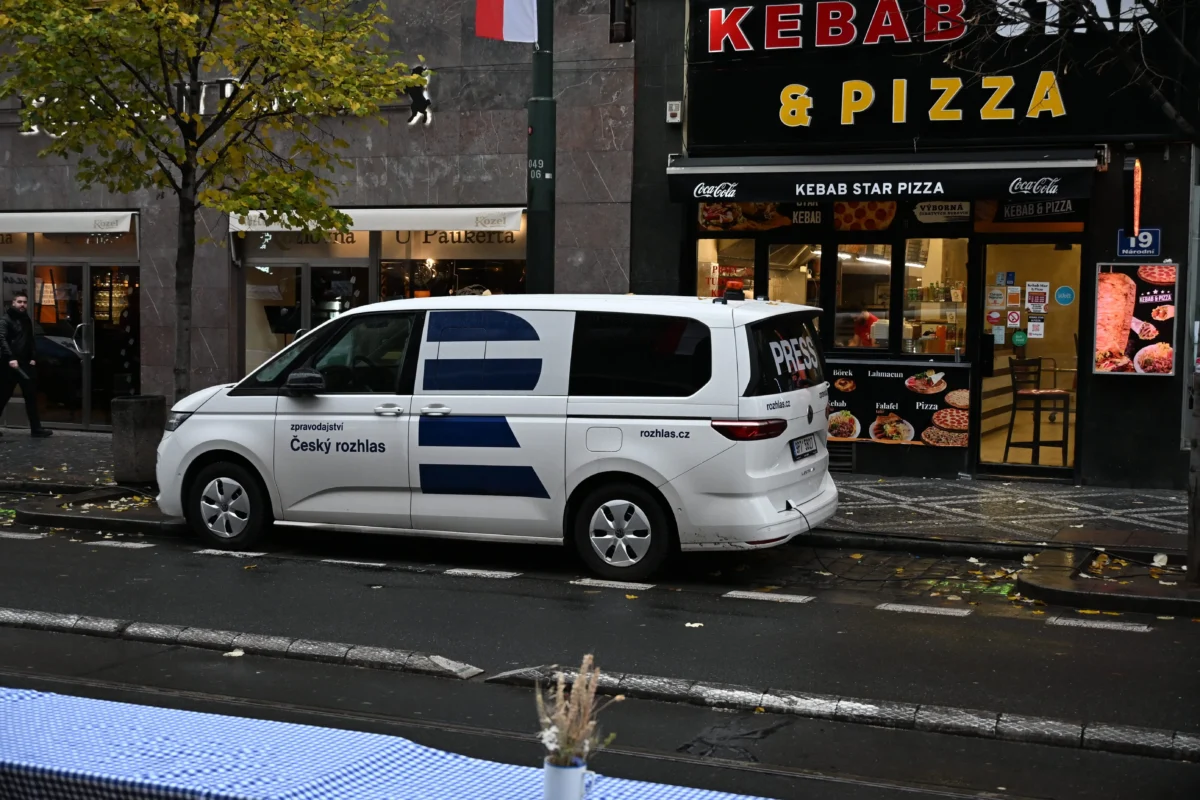 shutterstock_2706957119 1 | TVGURU.cz White Czech Radio van parked on a city street in front of a kebab/pizza shop, with a blue checked tablecloth in the foreground.