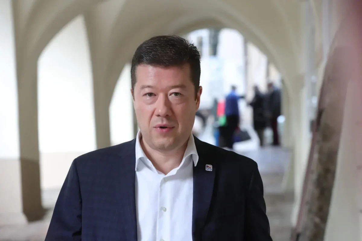 Man in a dark suit and white shirt walks toward the camera in a bright arched hallway; pin on lapel flourish act as SPD badge.
